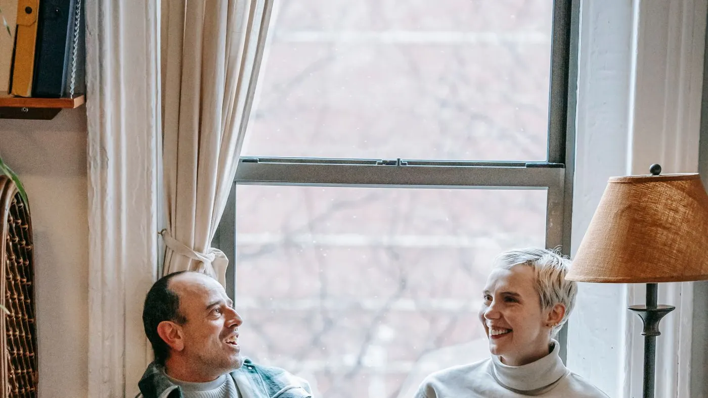 Pareja de mediana edad sonriendo y conversando junto a una ventana en la sala de su casa: imagen para la guía del potenciador sexual masculino