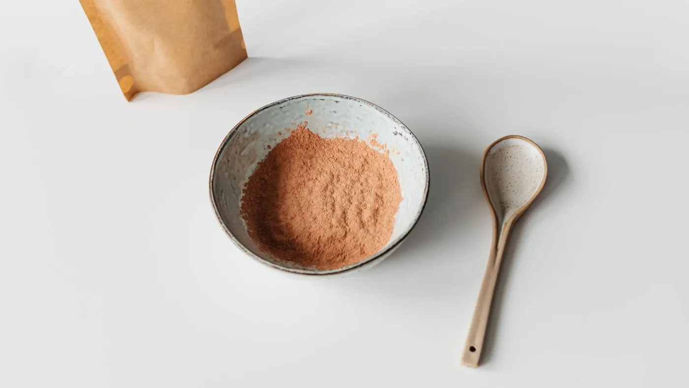 Polvo beige en un tazón de cerámica con cuchara de madera junto a una bolsa de papel: imagen para la guía de la maca en polvo